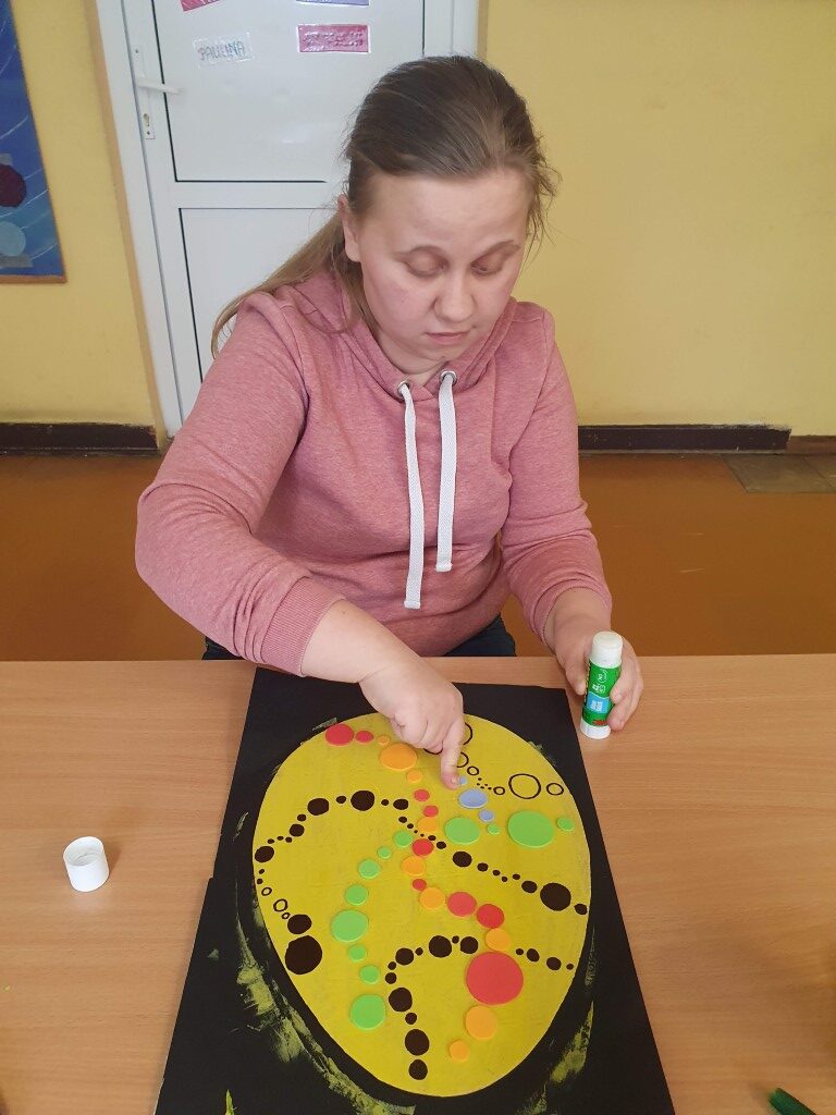 Uczennica przykleja do tekturowej pisanki pomalowanej na żółto kolejne kolorowe kropki.
