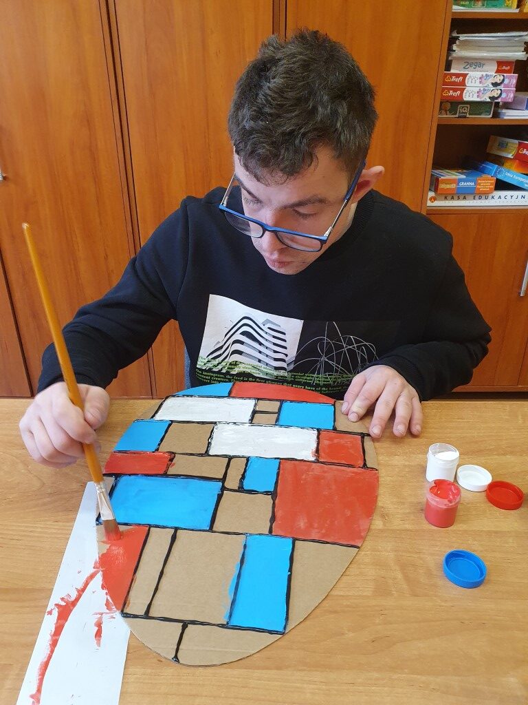 Uczeń maluje farbami pisankę z tektury podzieloną czarną grubą linią na geometryczne kształty. Kształty mają różne kolory: biały, czerwony, niebieski.