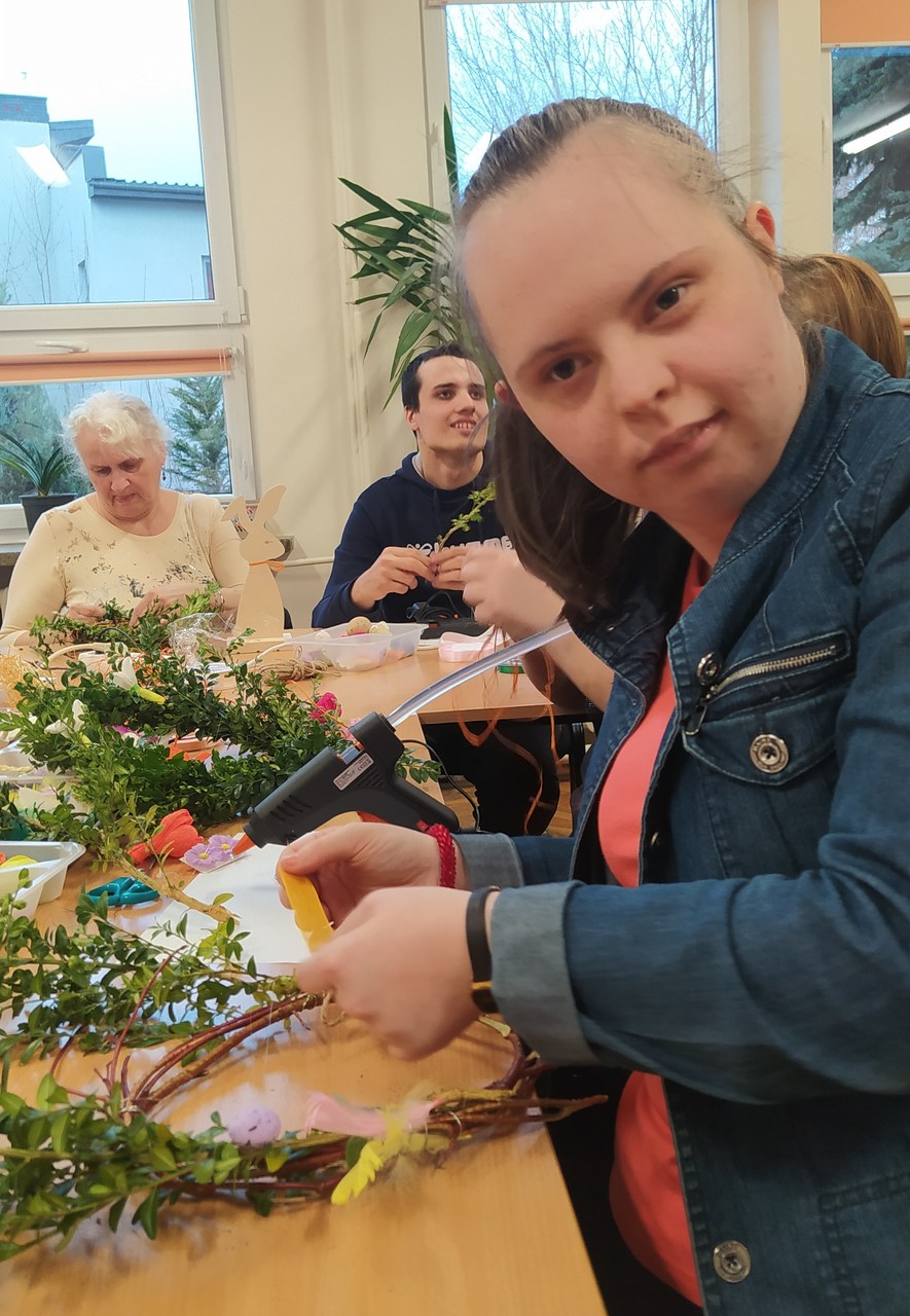 Uśmiechnięta uczennica ozdabia wianek kokardą obwiązując go nią z jednej strony.
