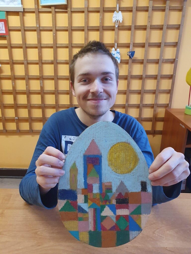 Uczeń prezentuje swoją tekturową pisankę pomalowaną pastelowymi kredkami w kolorowe figury tworzące zamek. Nad zamkiem świeci duże okrągłe słońce na niebieskim niebie.
