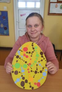 Uczennica prezentuje tekturową pisankę pomalowaną na żółto, na której kolorowe kropki tworzą różne wzory.