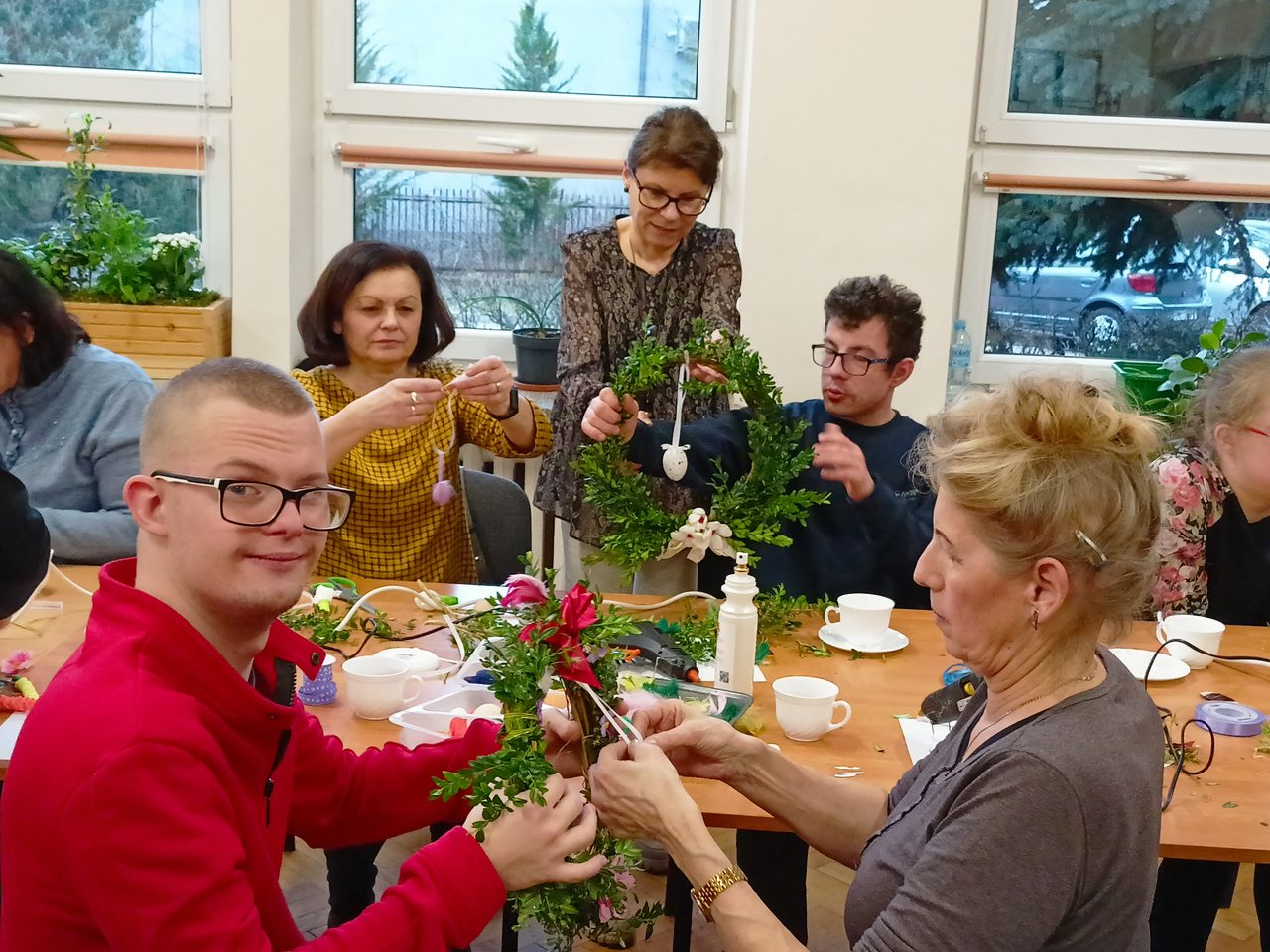 Na pierwszym planie uczeń trzyma wianek a jego mama wywiązuje kokardę. Na drugim planie pani dyrektor z uczniem ozdabia wianek. Mama ucznia doczepia kokardkę do pisanki.
