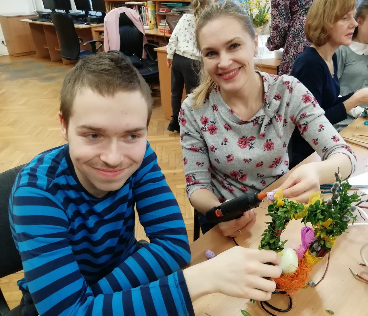 Uczeń przytrzymuje wianek a nauczycielka przykleja do wianka pisankę klejem na gorąco.