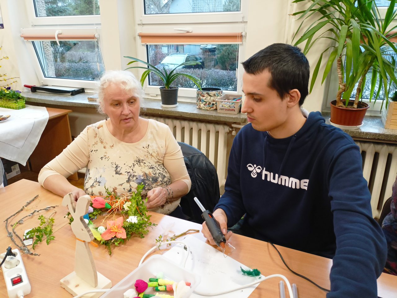 Uczeń trzyma pistolet na gorący klej a jego mama trzyma piękny duży wianek. Oboje zastanawiają się, jak mogą jeszcze ozdobić swoją pracę.
