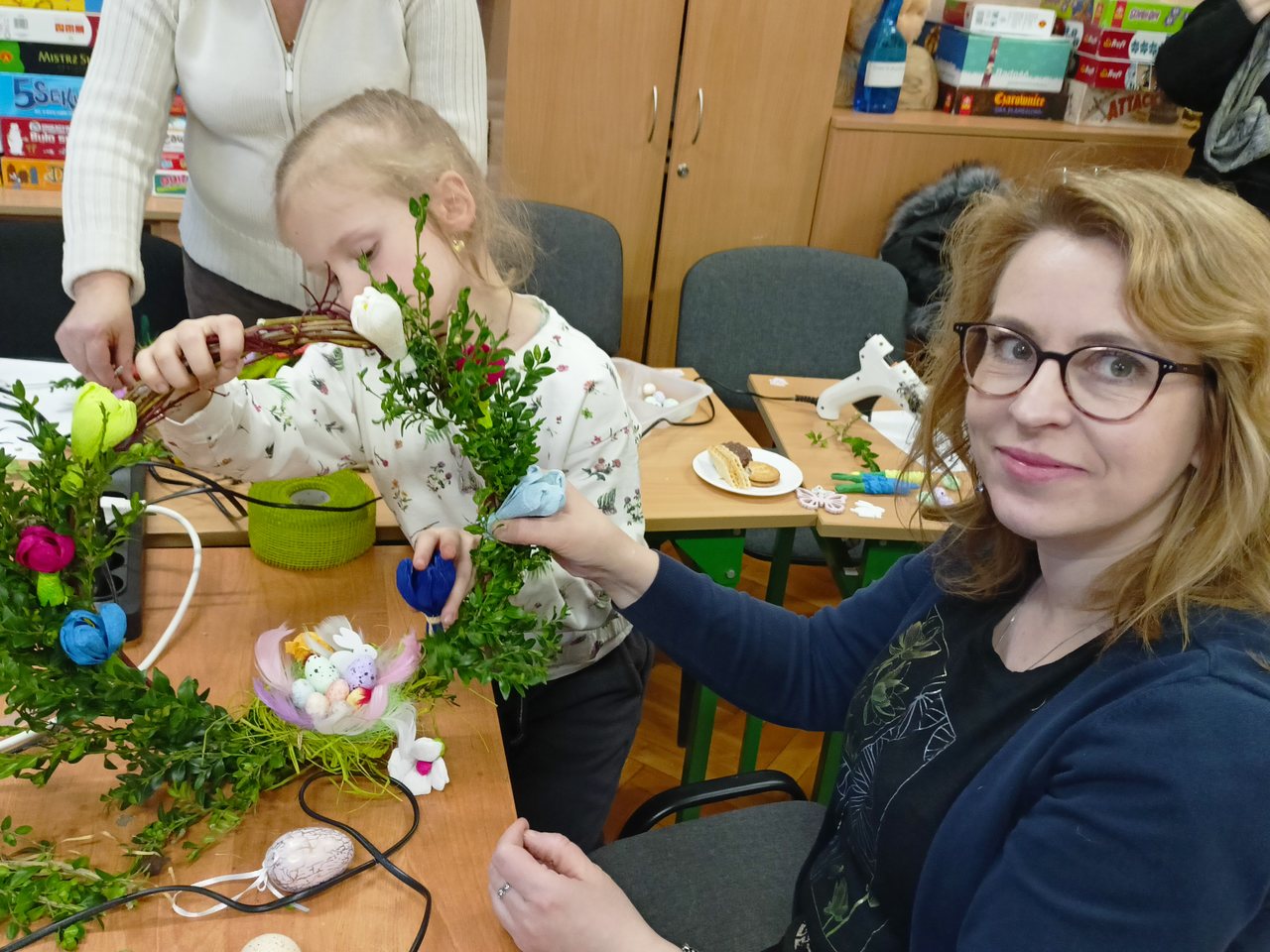 Ośmioletnia siostrzyczka uczennicy przyczepia kwiatek do wianka, który przytrzymuje jej mama.