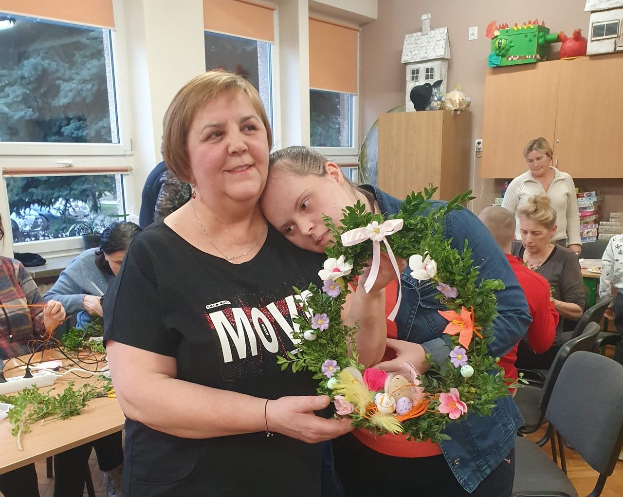 Uczennica przytula się do mamy. Obie trzymają i prezentują swój piękny wianek ozdobiony bukszpanem i kwiatami. Na dole wianka gniazdko z piórkami i kolorowymi pisankami. Na górze wianka wywiązana kokarda.