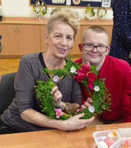 Uczeń siedzi z mamą za stołem. Mama prezentuje wspólnie wykonany wianek ozdobiony bukszpanem, małymi kwiatkami i dużą czerwoną kokardą. Oboje uśmiechają się.