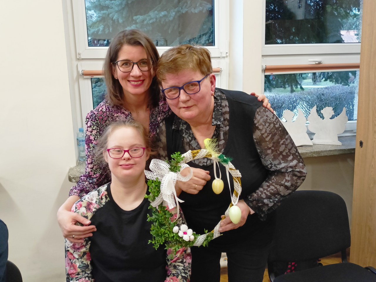 Uczennica siedzi za stołem a obok niej stoi jej mama i prezentuje wykonany wianek. Za uczennicą stoi jej wychowawczyni. Wszystkie osoby są do siebie przytulone i uśmiechają się.