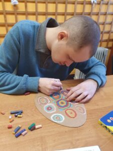 Uczeń maluje pastelowymi kredkami kartonową pisankę w kolorowe okręgi wpisane w kwadraty.