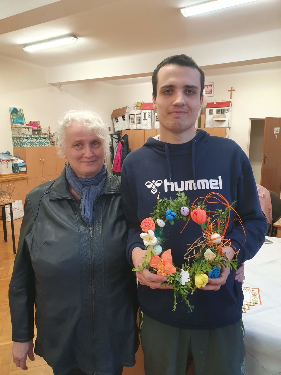 Uczeń stoi z mamą i prezentuje piękny wianek, który wykonali wspólnie. Ozdobili go bukszpanem oraz gęsto kolorowymi kwiatkami na zmianę z pisankami.