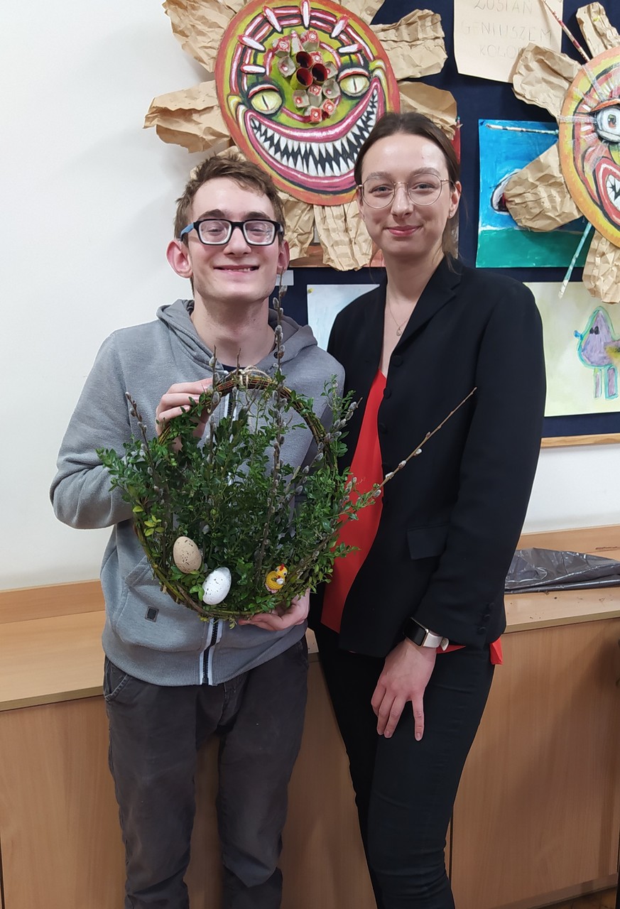 Uczeń stoi z siostrą i prezentuje wykonany wspólnie przez nich wianek ozdobiony dużą ilością bukszpanu, pisankami i kurczaczkiem. Oboje uśmiechają się.