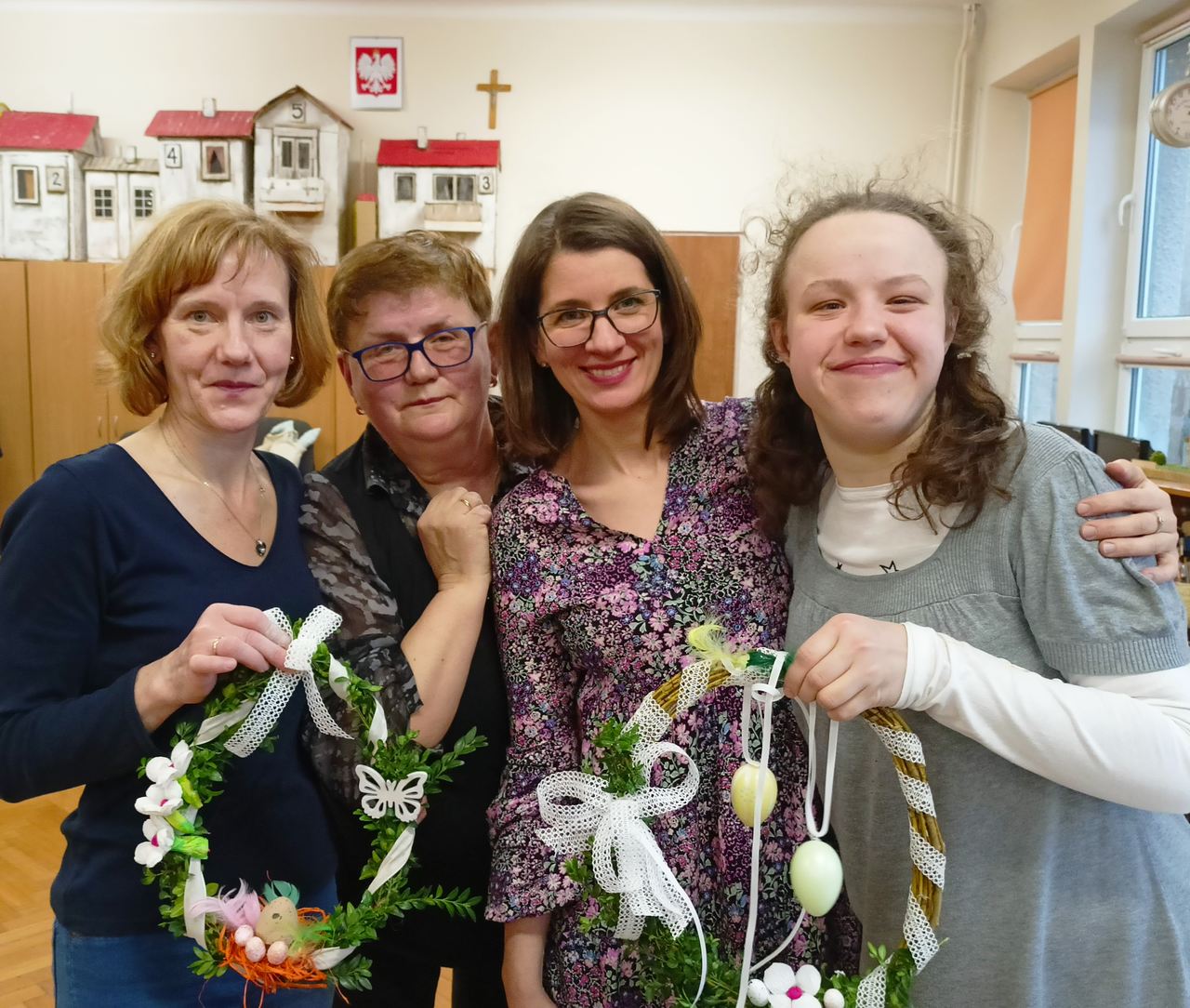 Przytulone stoją 4 kobiety: 2 mamy, wychowawczyni i jej uczennica. Jedna z mam i uczennica trzymają gotowe pięknie ozdobione wianki. Wszystkie osoby uśmiechają się.