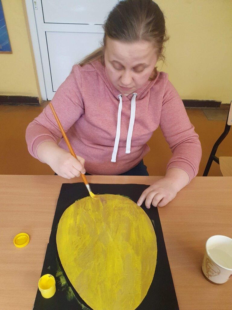 Uczennica maluje farbami kartonową pisankę na żółty kolor.