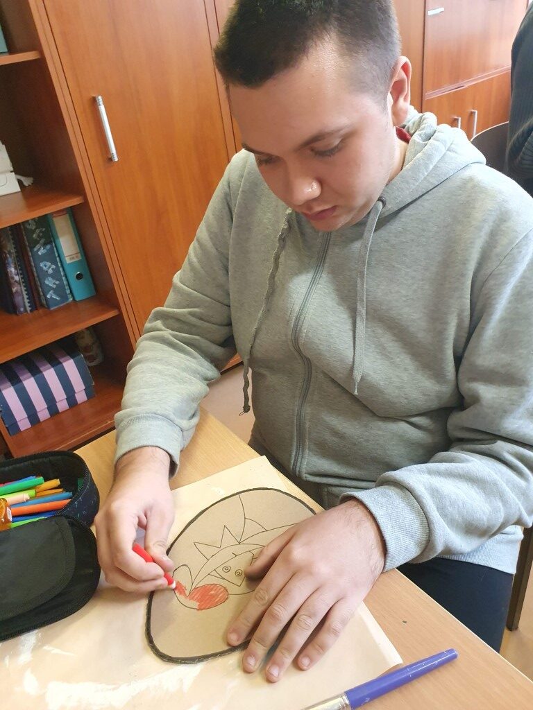 Uczeń na tekturowej pisance maluje kredkami kobietę w kapeluszu.