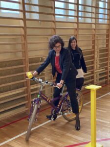 Na zdjęciu znajduje się Pani Dyrektor Zespołu Szkół nr16, która podczas jazdy na rowerze wykonuje jedno z zadań wymaganych na torze sprawnościowym w sali gimnastycznej.
