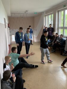 Na zdjęciu widoczne są grupy uczestników siedzących po lewej i po prawej stronie korytarza szkolnego, czekających na poszczególne konkurencje turnieju.