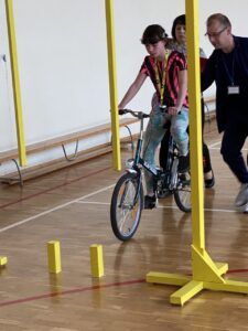Na zdjęciu widoczna jest uczestniczka turnieju jadąca na rowerze, pokonująca przeszkody na torze sprawnościowym, po prawej stronie wspierana przez sędziego.