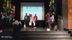 Na zdjęciu widocznych jest w oddali troje zwycięzców z nagrodami pozujących z głównym organizatorem na scenie auli szkolnej.