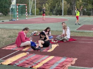 Grupa uczniów siedzi na kocach, w tle inni uczniowie grają w piłkę nożną na boisku szkolnym