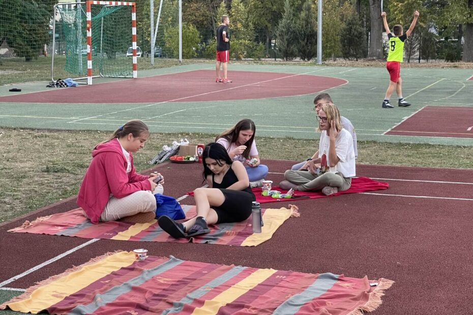 Grupa uczniów siedzi na kocach, w tle inni uczniowie grają w piłkę nożną na boisku szkolnym