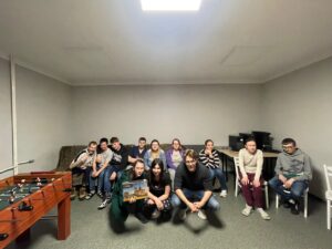 Uczestnicy siedzą w tym samym pomieszczeniu, tym razem w jaśniejszym oświetleniu. Kilka osób klęczy z przodu, trzymając pudełko z modelem katedry Notre-Dame de Paris – nagrodą za wygrany quiz. Na przodzie grupy znajduje się wychowawca ubrany na czarno. Reszta grupy siedzi na kanapie i krzesłach, a w tle widoczne są komputery ustawione na biurkach.