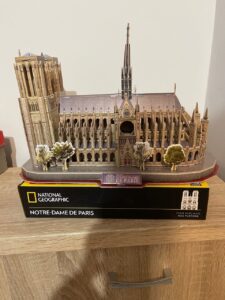 Model katedry Notre-Dame de Paris wykonany z puzzli 3D od National Geographic stoi na drewnianym meblu. Budowla jest bardzo szczegółowa – widoczne są wieże, rozeta, iglica oraz detale architektoniczne elewacji. Poniżej znajduje się pudełko z napisem „Your purchase has purpose,” podkreślającym dodatkową wartość zakupu.