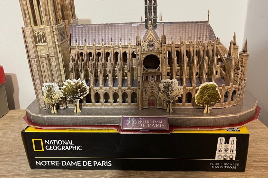 Model katedry Notre-Dame de Paris wykonany z puzzli 3D od National Geographic stoi na drewnianym meblu. Budowla jest bardzo szczegółowa – widoczne są wieże, rozeta, iglica oraz detale architektoniczne elewacji. Poniżej znajduje się pudełko z napisem „Your purchase has purpose,” podkreślającym dodatkową wartość zakupu.