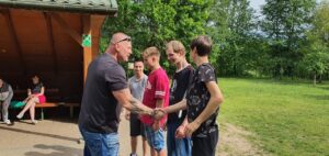 wychowawca uściskuje dłoń wychowanka ze zwycięskiej drużyny, w otoczeniu innych wychowanków.