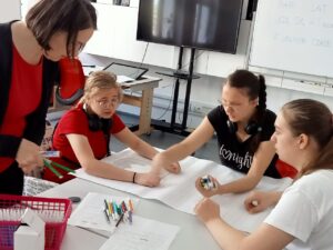 Nauczycielka nachylona nad trzema uczennicami siedzącymi przy stole udziela wyjaśnień jak wykonać zadanie używając kolorowych pisaków.