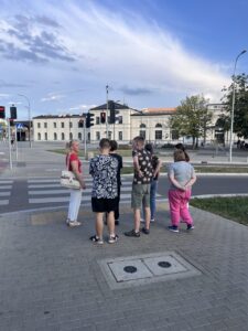 Wychowankowie wraz z panią Basią stoją przy przejściu dla pieszych z sygnalizacją świetlną, jest czerwone światło, co oznacza, że nie można w tej chwili przejść po pasach.