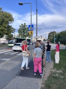 Wychowankowie wraz z panią Basią stoją przy przejściu dla pieszych. Pani Basia pokazuje wychowankom znak drogowy oznaczający, że w tym miejscu jest przejście dla pieszych