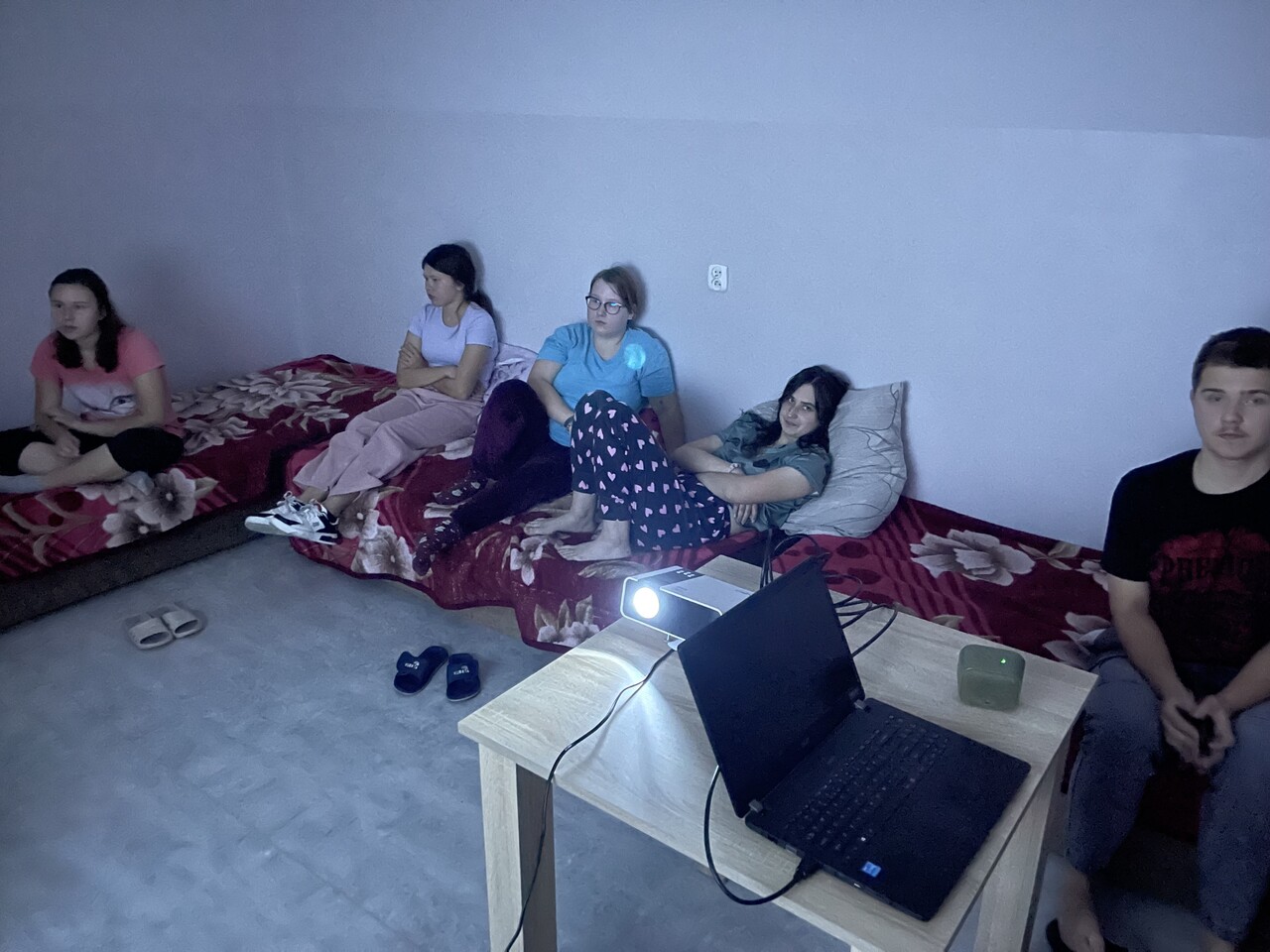 Na zdjęciu widać grupę siedmiu wychowanków rozłożonych na czerwonych łóżkach z bordowymi kocami w kwiatowe wzory. W pokoju panuje półmrok – jedyne światło pochodzi z projektora ustawionego na jasnym, drewnianym stoliku. Uczestnicy siedzą w luźnym półokręgu, skupieni na ekranie, trzymają w dłoniach poduszki i kubki z napojami.