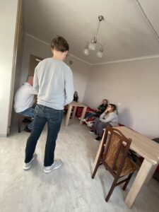 Dwóch wychowawców stoi w centrum pokoju, prowadząc dyskusję lub prezentację. Pozostali wychowankowie siedzą na łóżkach, słuchając i obserwując. Widoczna jest dynamika zajęć – uczestnicy aktywnie uczestniczą w rozmowie.