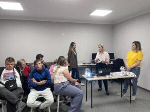 Wychowankowie siedzą na krzesłach, Panie prowadzące prowadzą wykład.
