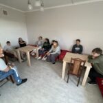 Wszyscy uczestnicy zajęć siedzą w pokoju – część na łóżkach, część na krzesłach przy stolikach. W centrum pomieszczenia widoczna jest przestrzeń do prowadzenia rozmów. Grupa tworzy krąg, co sprzyja otwartej dyskusji i wymianie myśli.