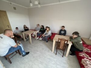 Wszyscy uczestnicy zajęć siedzą w pokoju – część na łóżkach, część na krzesłach przy stolikach. W centrum pomieszczenia widoczna jest przestrzeń do prowadzenia rozmów. Grupa tworzy krąg, co sprzyja otwartej dyskusji i wymianie myśli.