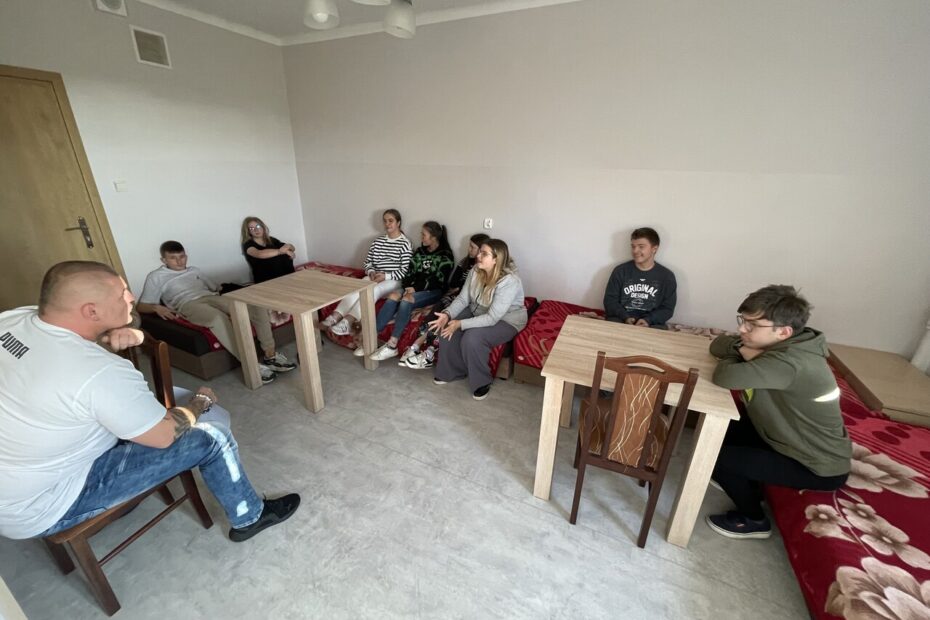 Wszyscy uczestnicy zajęć siedzą w pokoju – część na łóżkach, część na krzesłach przy stolikach. W centrum pomieszczenia widoczna jest przestrzeń do prowadzenia rozmów. Grupa tworzy krąg, co sprzyja otwartej dyskusji i wymianie myśli.