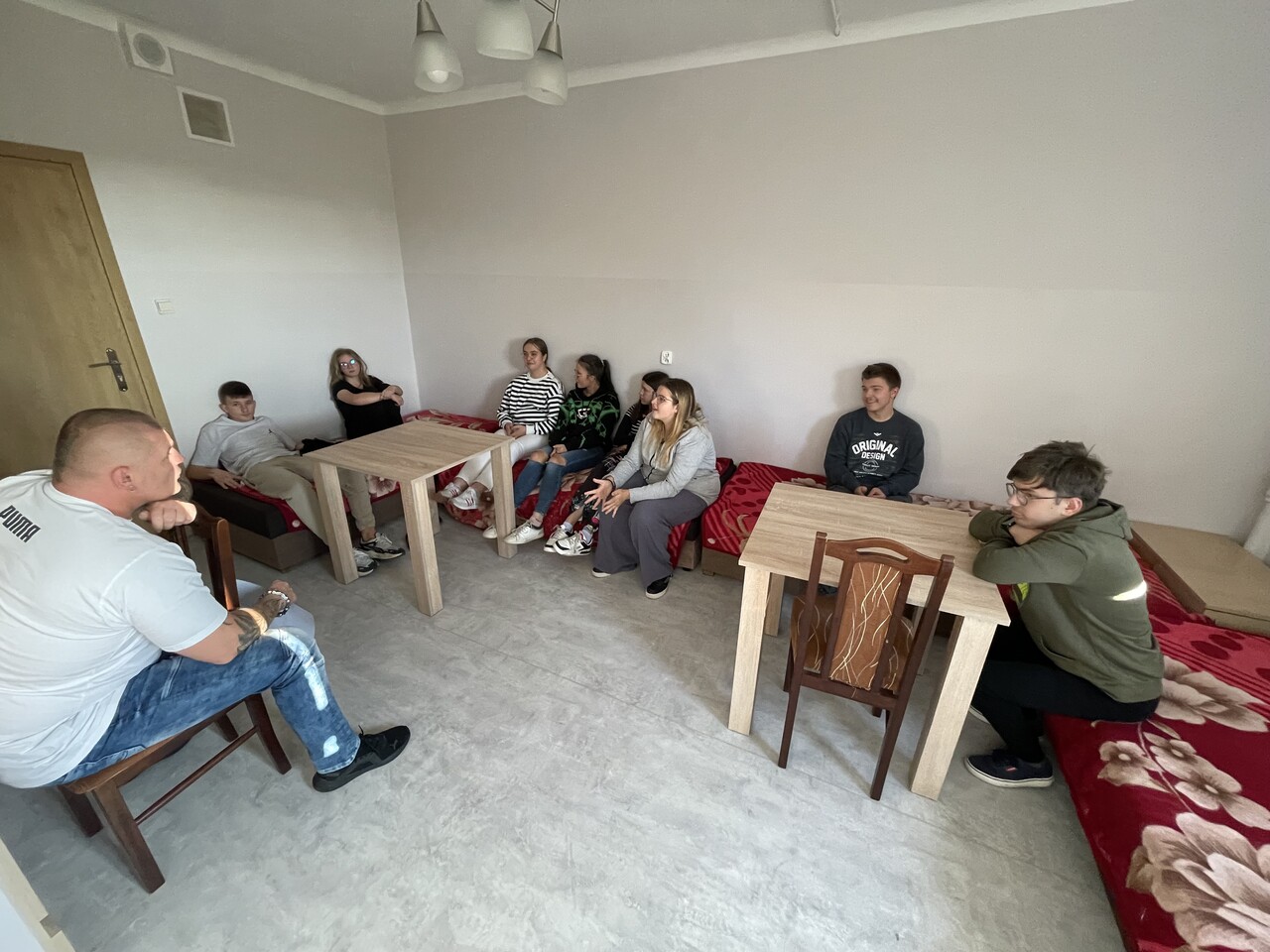 Wszyscy uczestnicy zajęć siedzą w pokoju – część na łóżkach, część na krzesłach przy stolikach. W centrum pomieszczenia widoczna jest przestrzeń do prowadzenia rozmów. Grupa tworzy krąg, co sprzyja otwartej dyskusji i wymianie myśli.