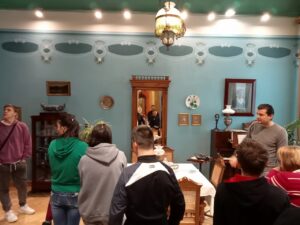 Dziewczęta i chłopcy słuchają opowieści przewodnika muzeum i oglądają piękne wnętrze salonu z początku XX wieku.
