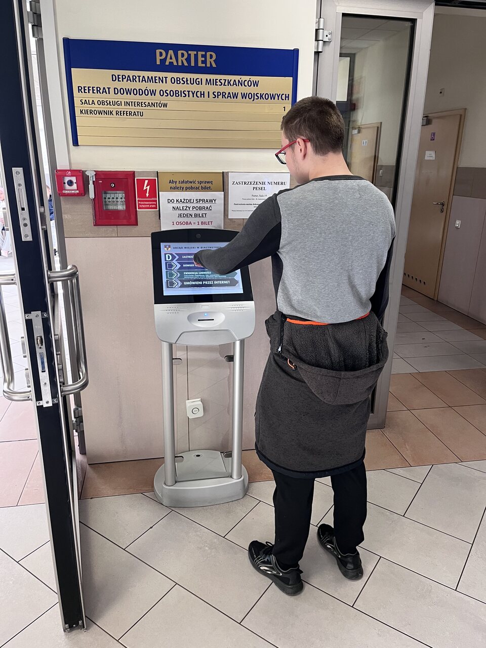 Wychowanek stoi przy monitorze, gdzie wybiera, którą sprawę urzędową chce załatwić.
