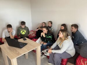 Grupa ośmiorga wychowanków siedzi na czerwonych łóżkach w jasnym pokoju internatu, skupiona na oglądaniu prezentacji wyświetlanej na laptopie. Wszyscy słuchają z uwagą, część osób ma zamyślone miny. Na pierwszym planie widoczny jest drewniany stolik z czarnym laptopem.