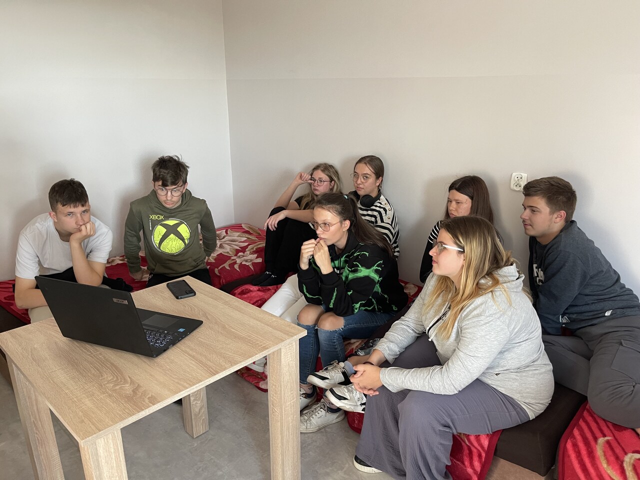 Grupa ośmiorga wychowanków siedzi na czerwonych łóżkach w jasnym pokoju internatu, skupiona na oglądaniu prezentacji wyświetlanej na laptopie. Wszyscy słuchają z uwagą, część osób ma zamyślone miny. Na pierwszym planie widoczny jest drewniany stolik z czarnym laptopem.