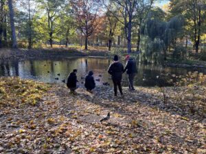 Wychowankowie stoją nad stawem i przyglądają się pływającym w stawie kaczkom.
