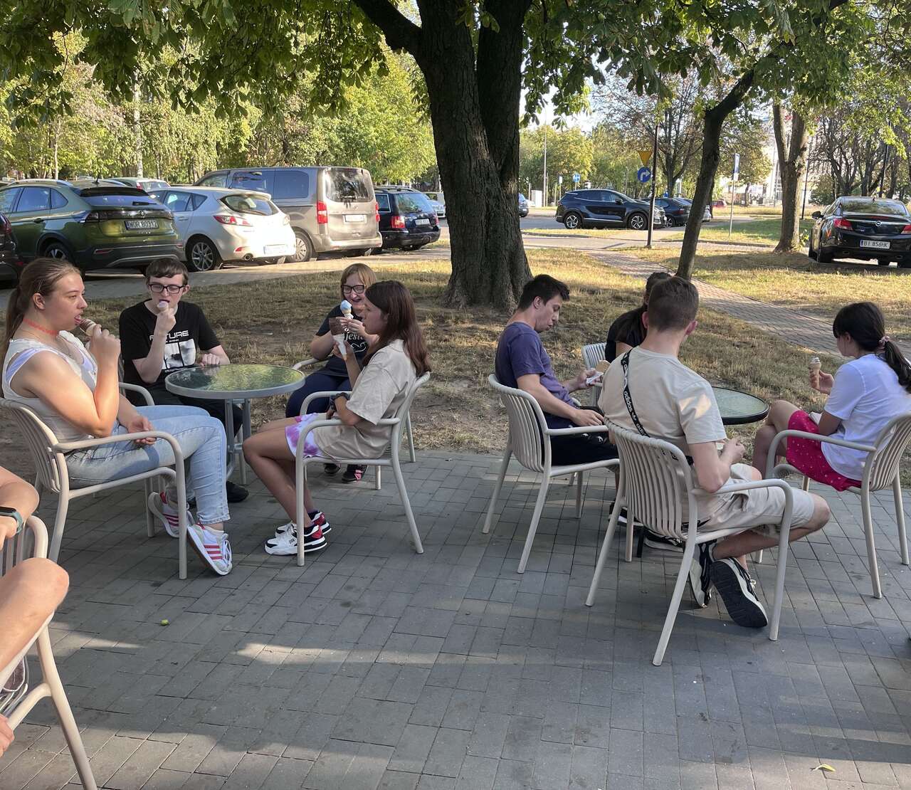 Młodzież siedzi przy stolikach jedząc lody.