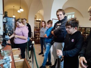 Młodzież znajduje się w dawnej aptece Pałacu Branickich. Jedna z osób będących w grupie obsługuje stare narzędzia aptekarskie.
