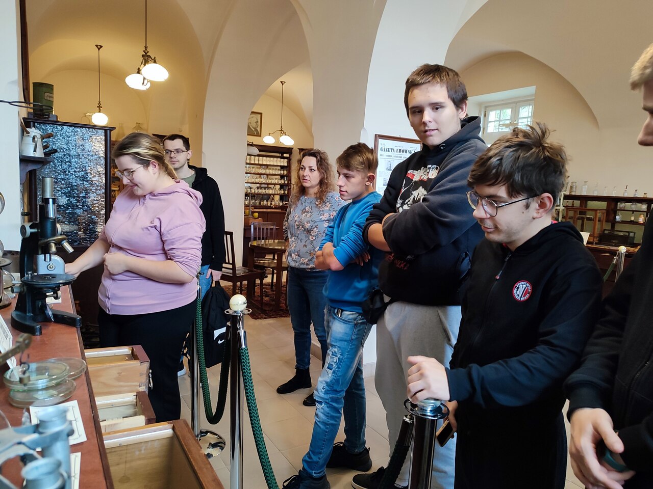Młodzież znajduje się w dawnej aptece Pałacu Branickich. Jedna z osób będących w grupie obsługuje stare narzędzia aptekarskie.
