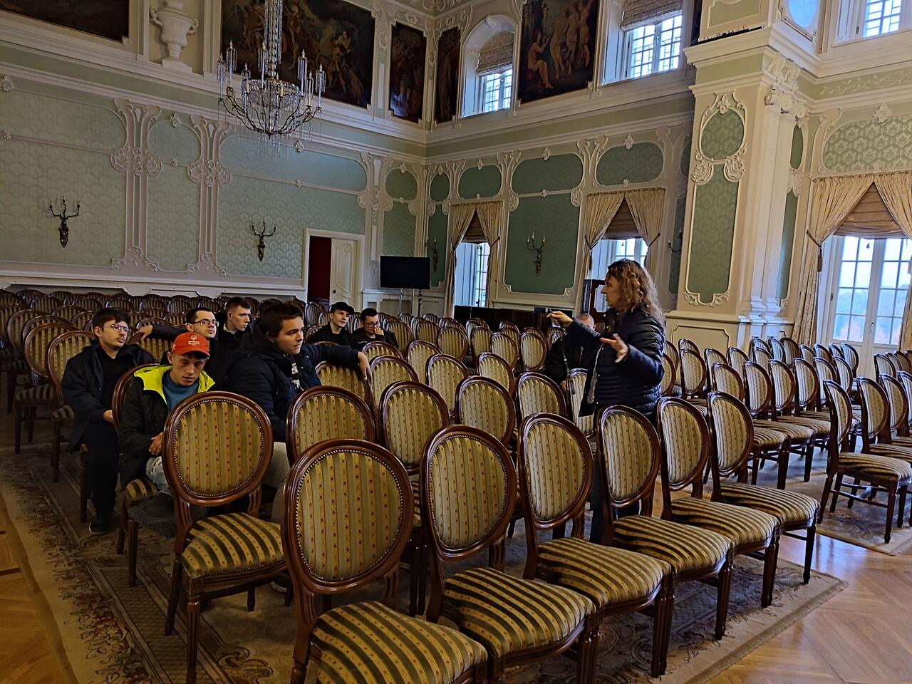 Zdjęcie przedstawia grupę młodzieży w Sali balowej Pałacu Branickich. Grupa z zaciekawieniem słucha pani przewodnik.
