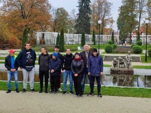 Grupa młodzieży wraz z opiekunami stoi obok niewielkiego stawu i rzeźb, otoczona jesiennymi drzewami i elegancką zielenią.
