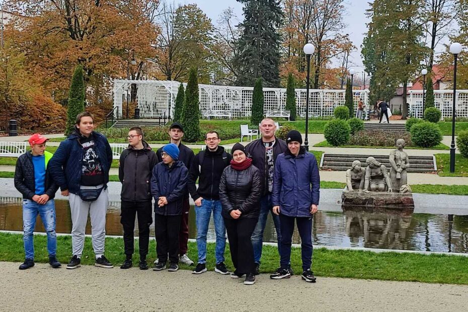 Grupa młodzieży wraz z opiekunami stoi obok niewielkiego stawu i rzeźb, otoczona jesiennymi drzewami i elegancką zielenią.