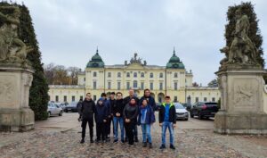 Grupa stoi na placu przed główną fasadą Pałacu Branickich, pomiędzy dwoma dużymi rzeźbami.